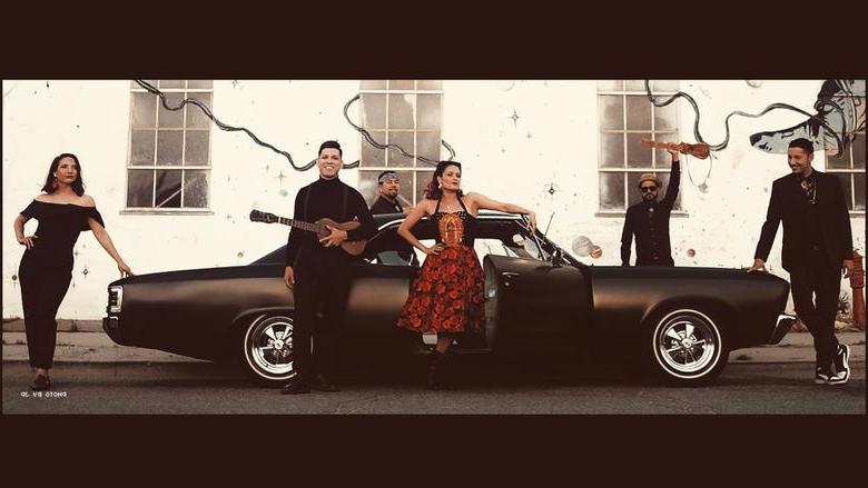 Members of a band hold instruments while standing around a black car